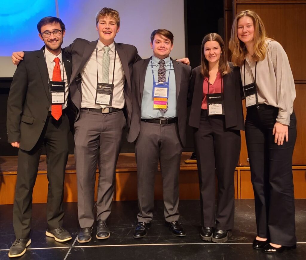 Five young adults in conference attire smiling and standing arm-in-arm on a stage, wearing name badges and lanyards, with a large screen in the background.