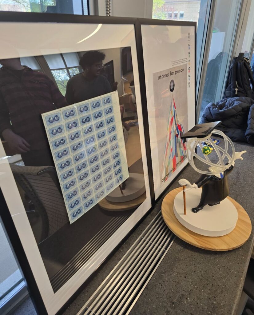 Framed display featuring a full sheet of commemorative nuclear-themed postage stamps and an "Atoms for Peace" poster next to a small figurine of a person in academic regalia holding a pointer, with atomic orbit rings and a model of Earth over their head, all on a round wooden base.