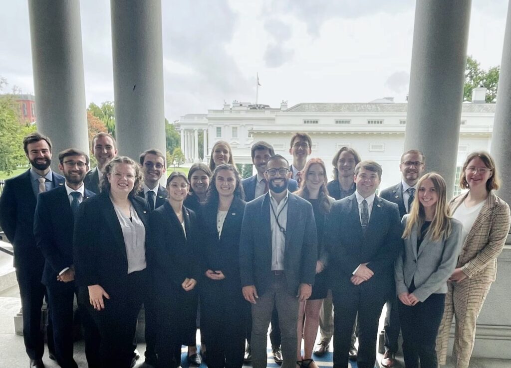 Group photo of professionally dressed individuals posing on the White House colonnade with the White House in the background, taken during an official visit or advocacy day.