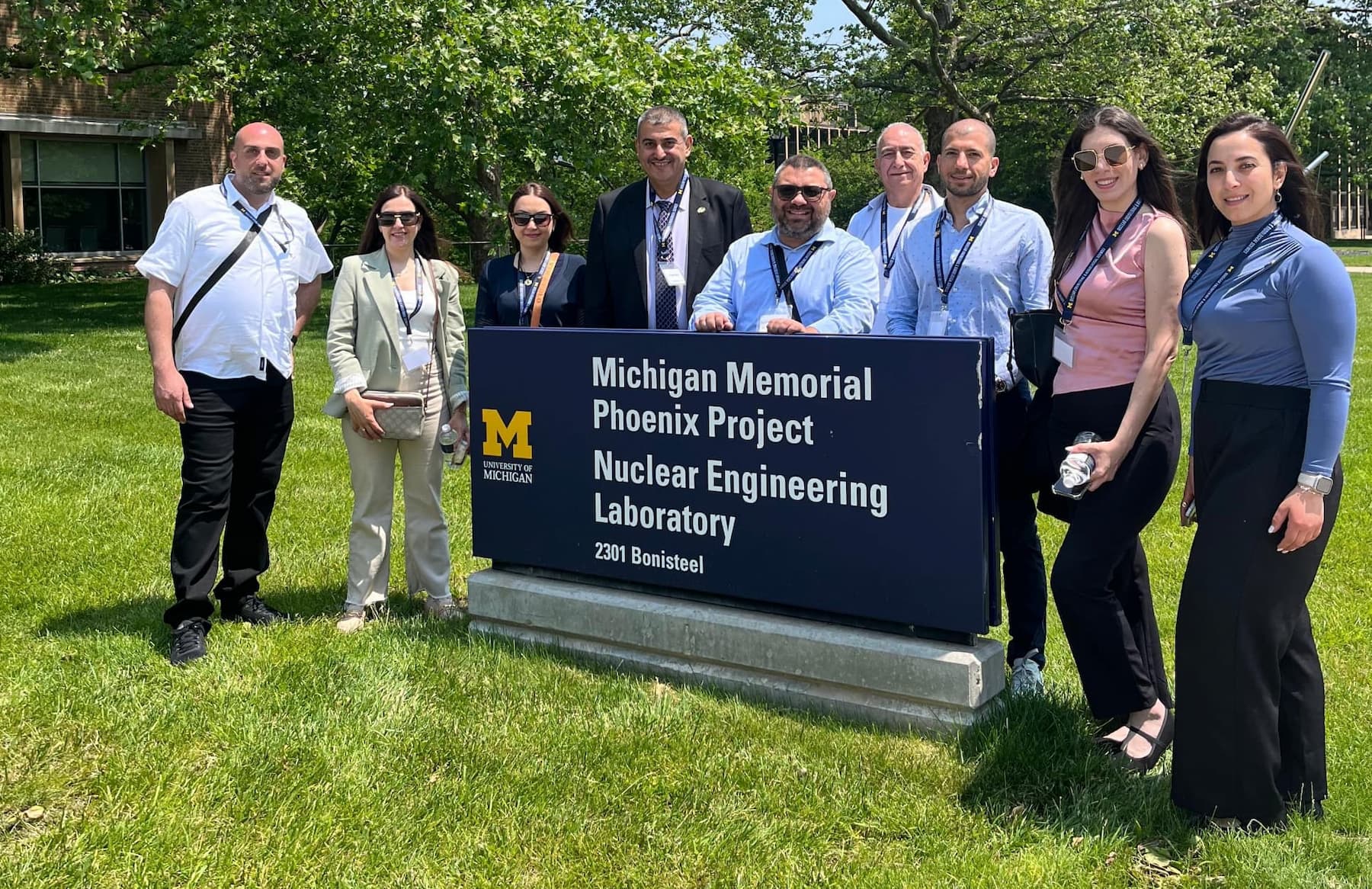 A group of nine people poses for a photo next to a blue University of Michigan sign that reads “Michigan Memorial Phoenix Project – Nuclear Engineering Laboratory – 2301 Bonisteel.” They are outdoors on a sunny day, standing on green grass with leafy trees in the background. Most individuals are wearing business casual attire with name badges on lanyards, suggesting they are visitors or participants in an organized tour or event.