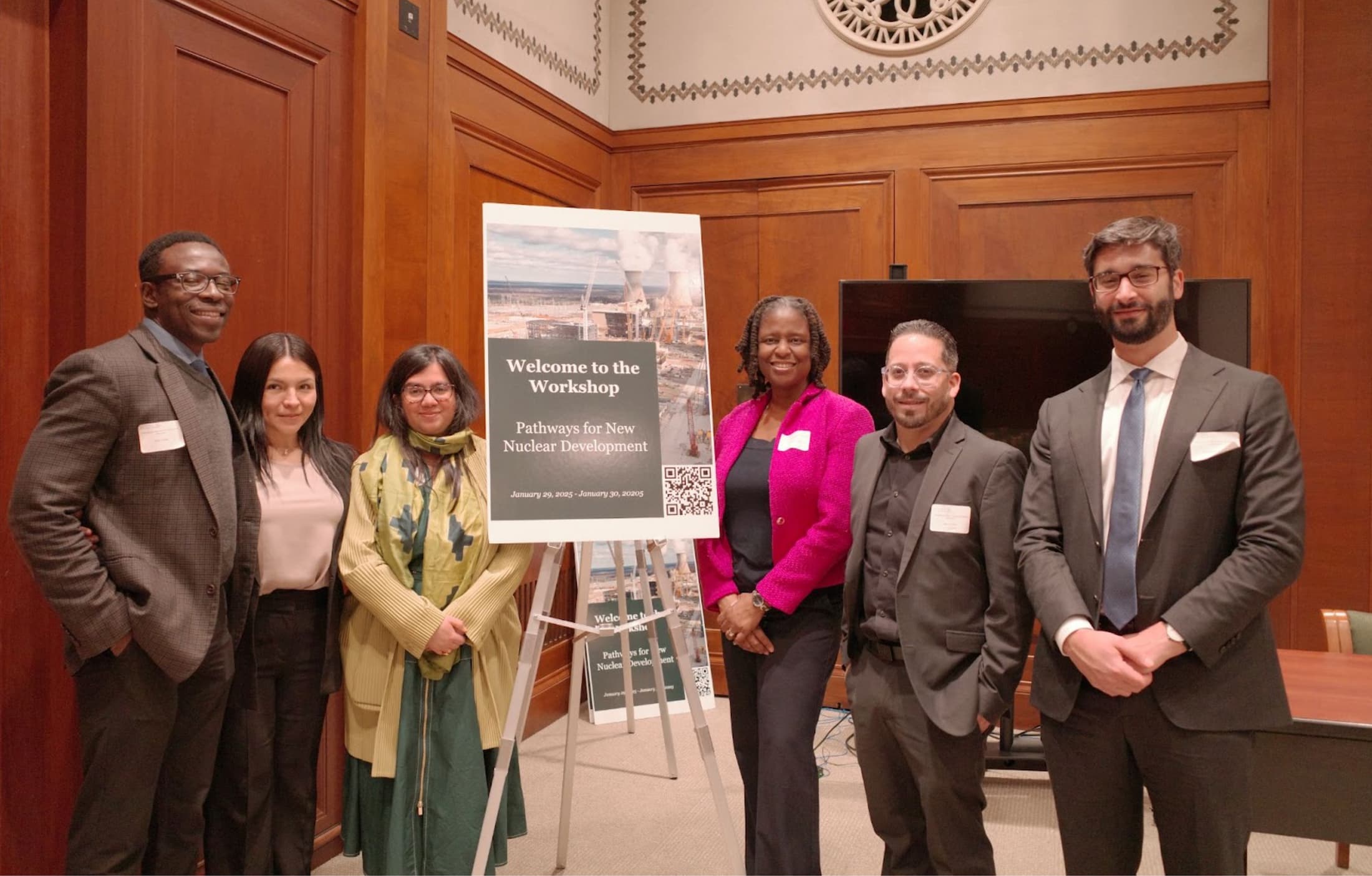Six people stand together smiling beside a sign that reads “Welcome to the Workshop: Pathways for New Nuclear Development, January 29, 2025.” The group is posed in a wood-paneled room with ornate details on the wall.